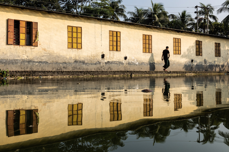 A man walks along a wall reflected in water in Barisal